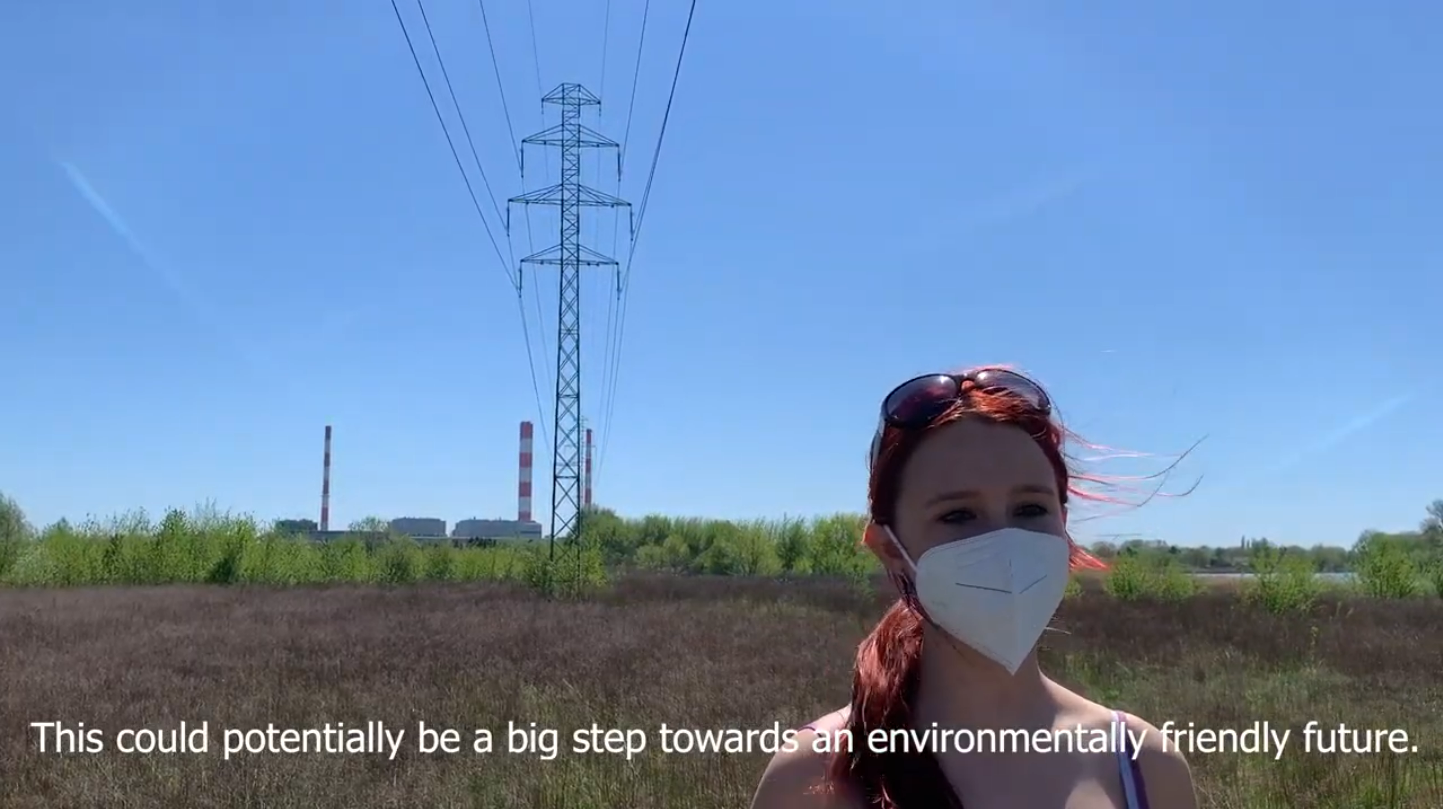A presenter stands in a field beneath power lines and industrial stacks while discussing environmentally friendly futures.