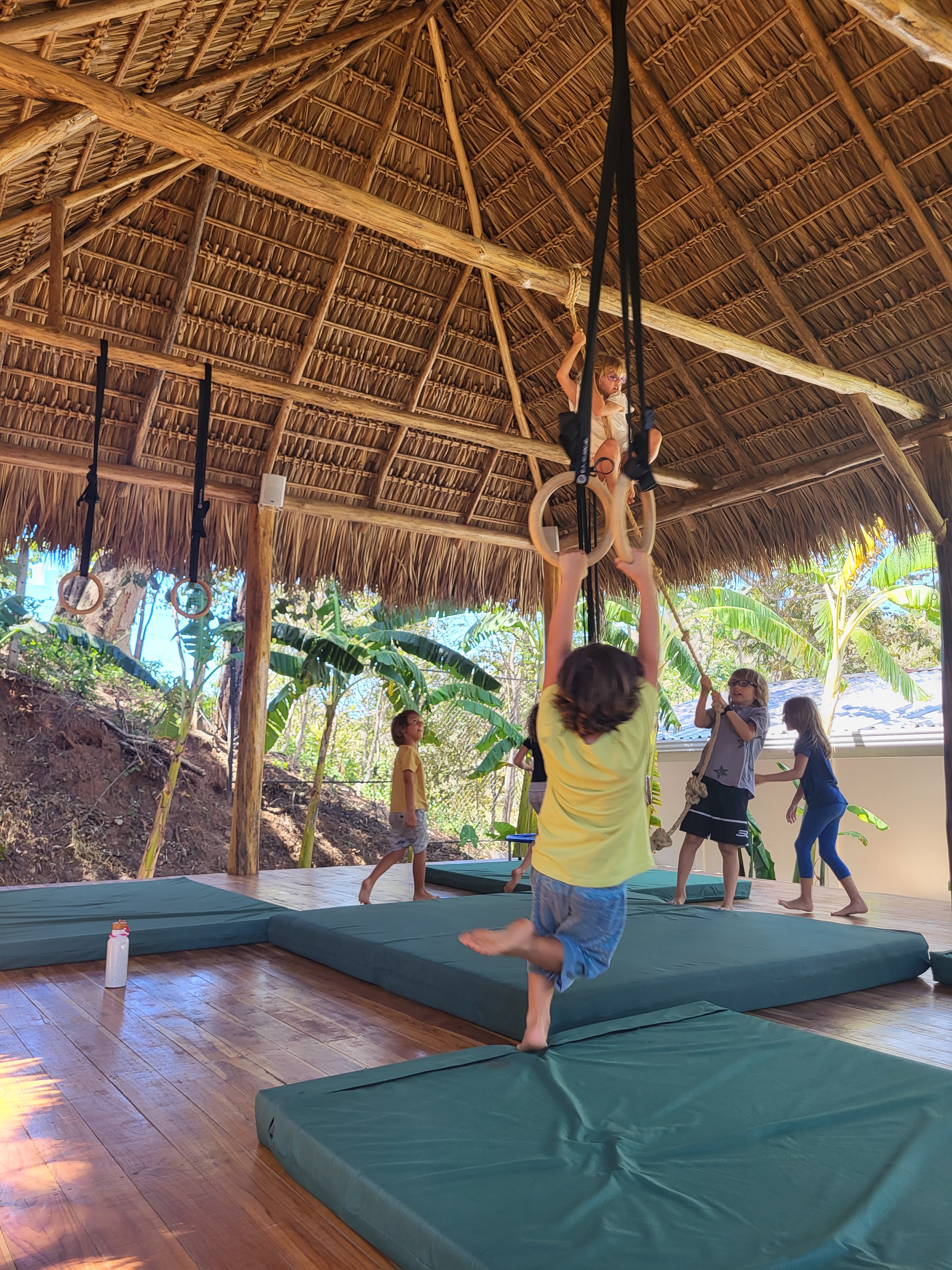 Primary school children playing on gymnastic rings inside an open-air thatched pavilion at NEO School Playa Grande.